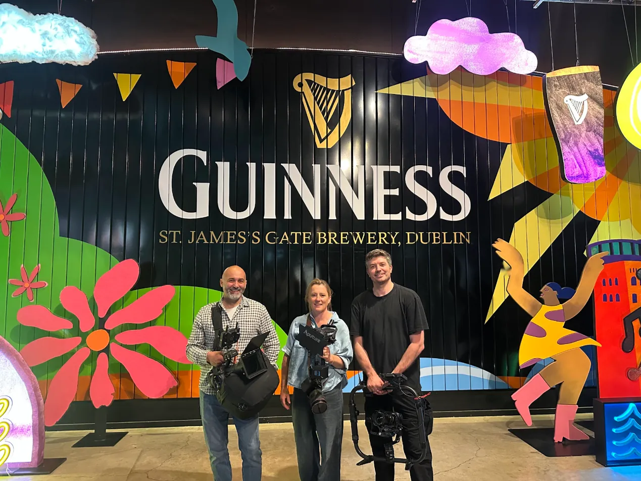 Emma with Blat and Dan from the crew, holding cameras and a stabiliser rig in front of the Guinness summer mural, with a painted harp, sun and dancing figure behind them.